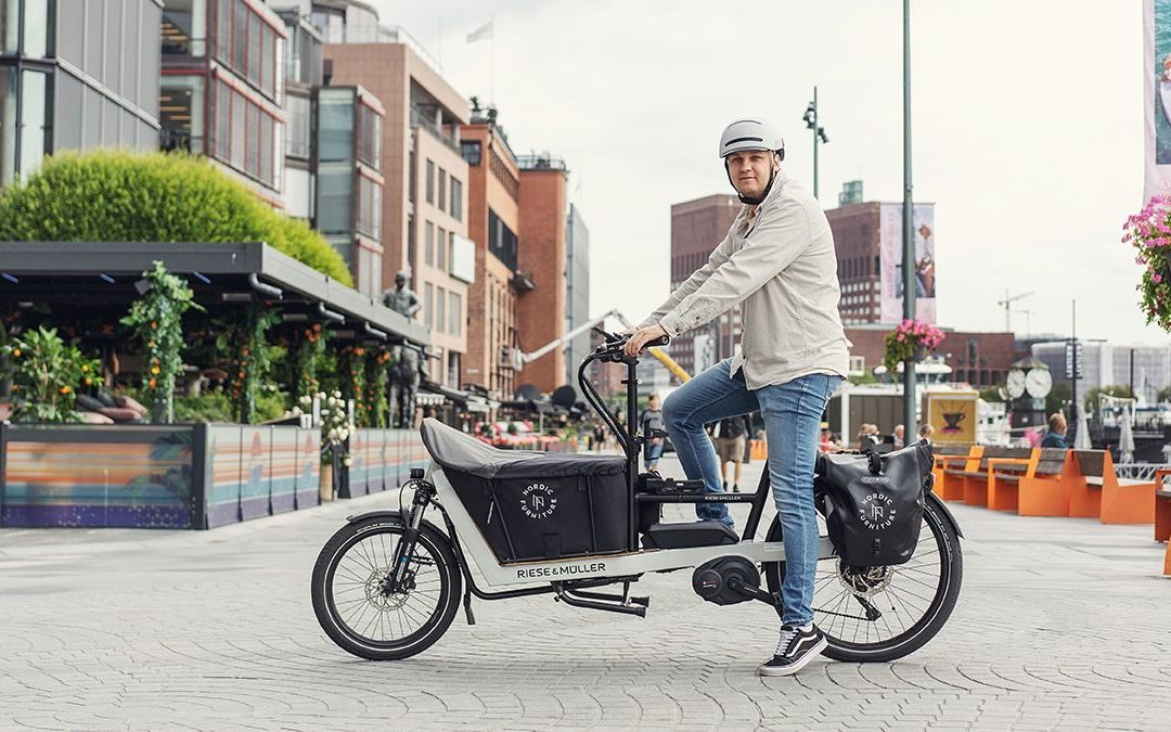 En person i hjelm sykler på en lastesykkel i en bygate. Bygninger og uteservering er synlig i bakgrunnen.