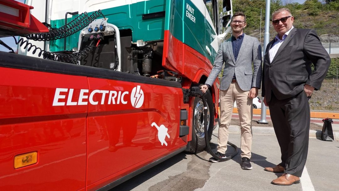 To menn står ved siden av en stor rød og hvit lastebil merket ELECTRIC, som eksemplifiserer en forgangsby for utslippsfri tungtransport. En mann holder en ladekabel koblet til lastebilen.
