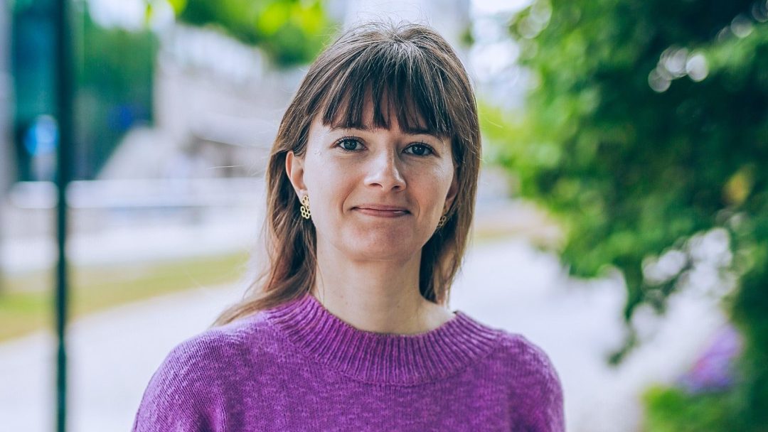 En kvinne med langt hår står utendørs, iført en lilla genser, og tenker på viktigheten av klimainvesteringer. Trær og en uskarp bakgrunn er synlige, og rammer inn hennes gjennomtenkte holdning i naturen.