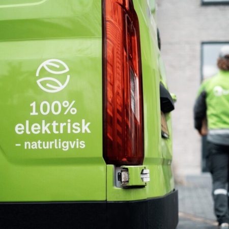 Grønt elektrisk kjøretøy med teksten 100% elektrisk - selvfølgelig, som legemliggjør moderne klimakrav. En person i en grønn uniform går bort, med den uskarpe bakgrunnen til en bygning som forsterker scenens dynamiske energi.