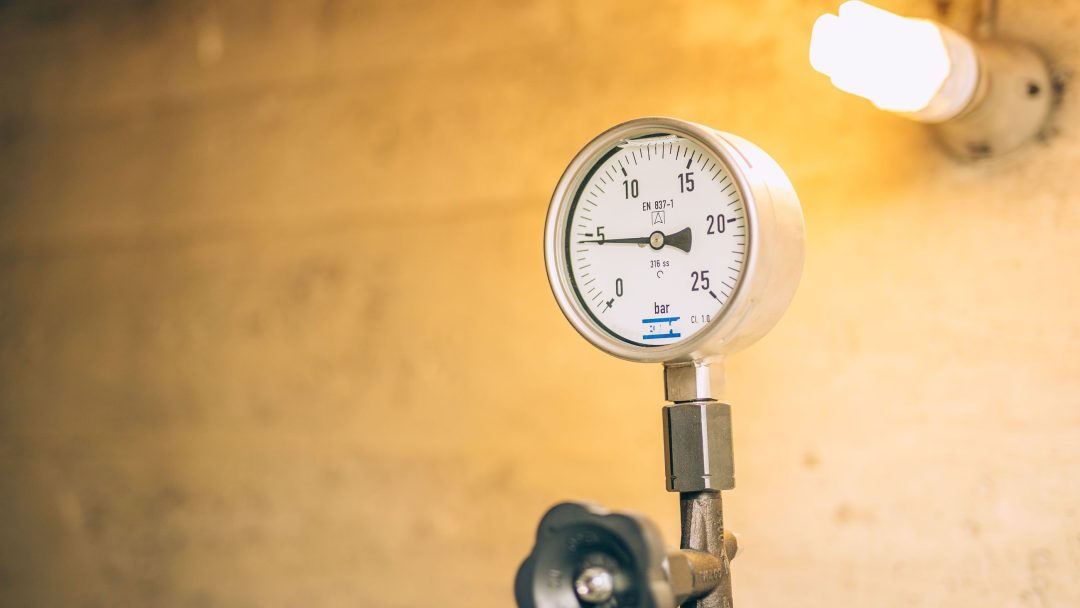 En trykkmåler montert på et rør mot en beige vegg, med en lyspære som lyser i bakgrunnen, hinter om pågående isoleringsarbeid for å øke effektiviteten.