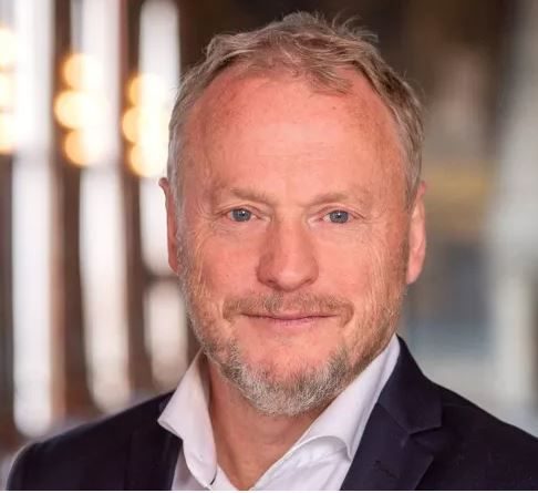 En mann med kort, lyst hår og skjegg, iført dress og hvit skjorte, smiler til kameraet. Den svakt opplyste bakgrunnen glitrer som prismer som danser i uskarpt lys.