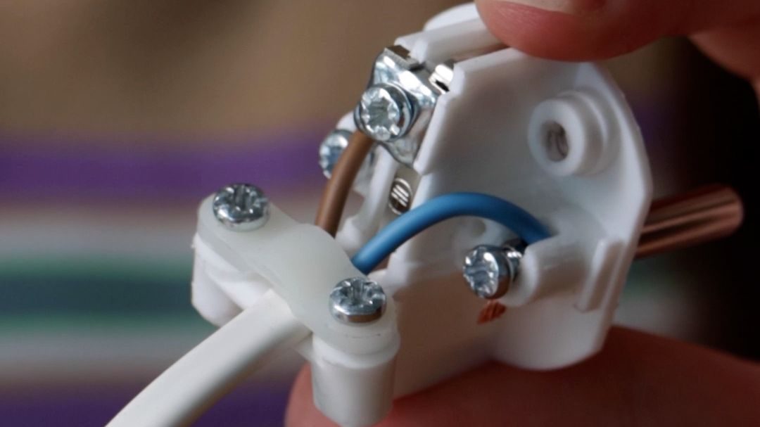 Nærbilde av en persons hånd som holder en hvit elektrisk plugg, eller støpsel. Pluggen har synlige ledninger koblet med skruer, som viser blå og brune ledninger.