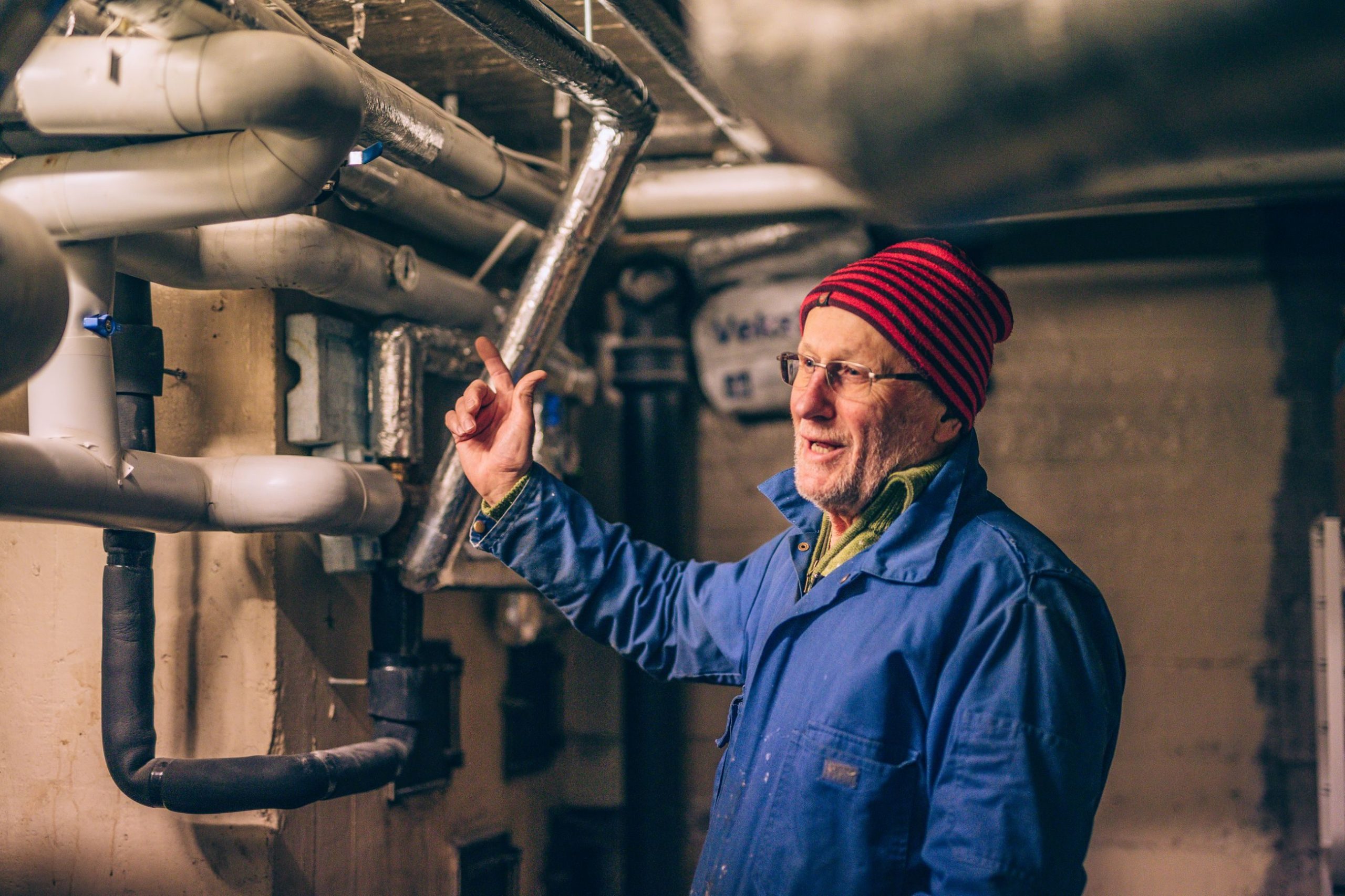 En eldre mann i blå frakk og rødstripet lue peker på komplekse rør i en kjeller, og forklarer energiordninger for en liten gruppe.