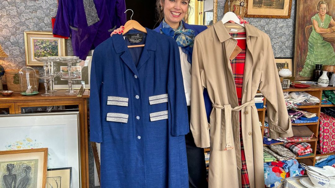 En kvinne står i et rom med en blå frakk og en beige trenchcoat, som minner om funn ved et loppemarked. Bakgrunnen har innrammede kunstverk og forskjellige gjenstander på et bord, noe som bidrar til den eklektiske sjarmen.