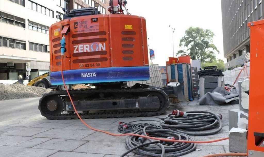 Et oransje anleggskjøretøy buldrer langs en bygate og avgir lave utslipp mens det manøvrerer rundt kabler på bakken. I bakgrunnen står bygninger og byggematerialer klare for fremgang.