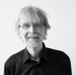 En mann med briller og skjegg smiler til kameraet, og utstråler en følelse av stille selvtillit som ligner på å presentere en feilfri synteserapport. Han har på seg en svart skjorte.