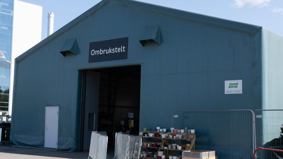 Et stort grønt telt merket Ombrukstelt, med åpen inngang, omgitt av metallbarrikader og et skilt på høyre vegg, gir et innblikk i bærekraftig livsstil med sitt utvalg av brukte møbler.