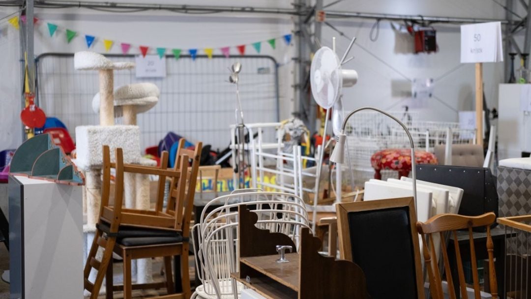 En rotete lagringsplass fylt med stablede stoler, et kattetre, en lampe og en vifte, sammen med diverse brukte møbler. Flerfarget bunting henger over.