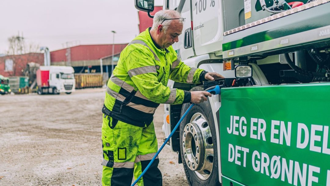 En mann i synlige klær kobler en kabel inn i et stort grønt kjøretøy, sannsynligvis en lastebil, med teksten JEG ER EN DEL AV DET GRØNNE... på et uteområde.