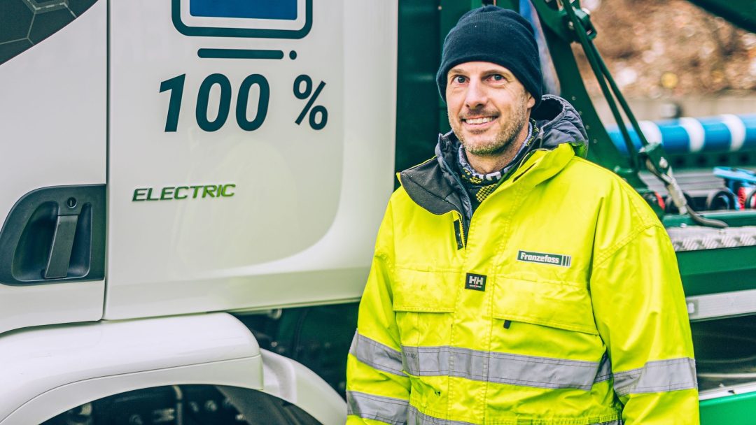 En person i en knallgul jakke står foran en hvit elektrisk lastebil med 100% skrevet på.