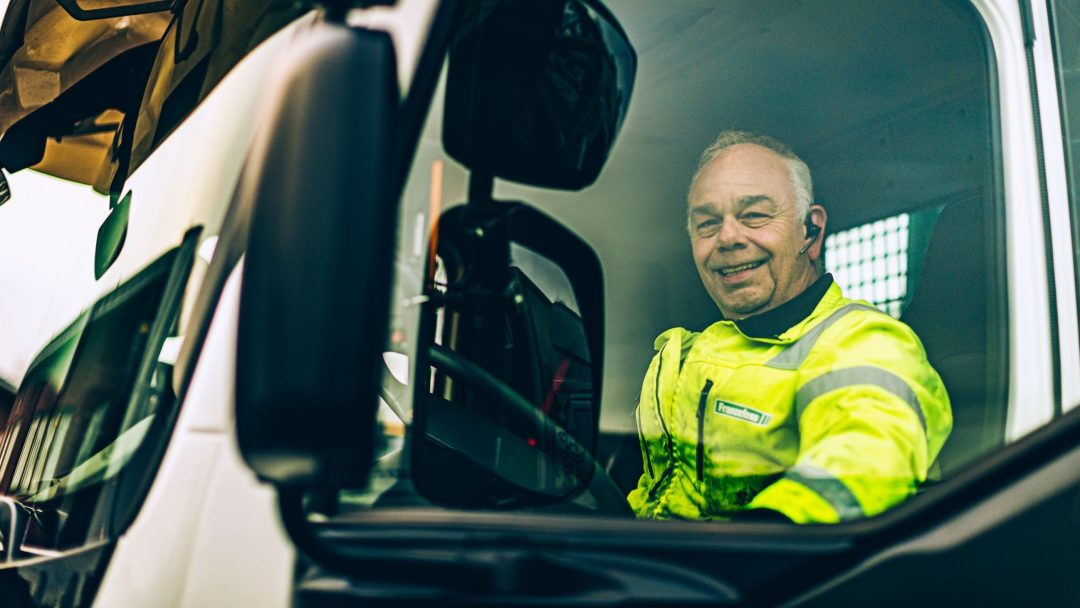 Mann i synlig jakke sitter i førersetet på en lastebil, smiler og ser ut av vinduet.