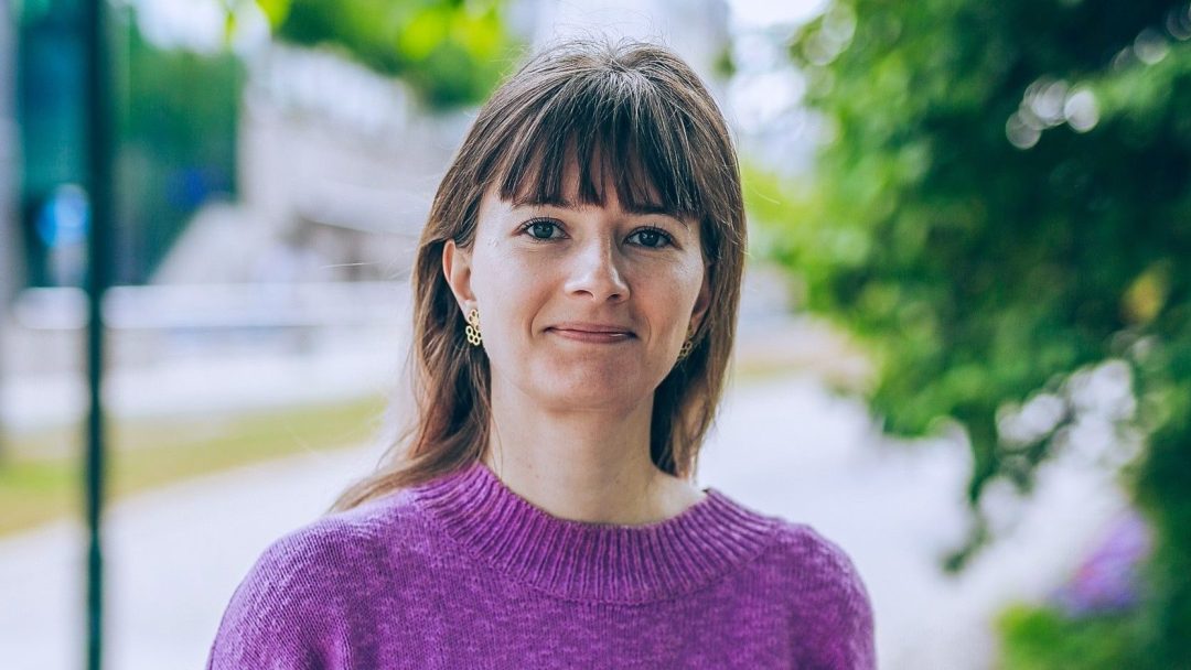 En person med langt hår og øredobber har på seg en lilla genser og står utendørs nær grøntområder.