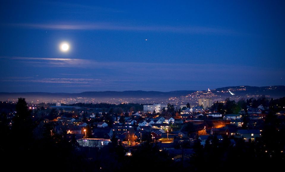 Et bybilde om natten med fullmåne på himmelen, der skimrende bylys lyser opp bygningene og gatene nedenfor, alt utfolder seg under et rolig bakteppe som subtilt hvisker av klimamålet. Horisonten avslører svake konturer av fjerne åser.