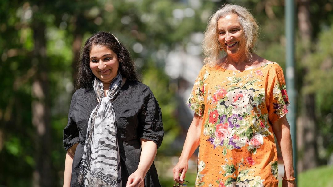 To kvinner som går utendørs på en solrik dag, kanskje på vei til jobb. Den ene har på seg en svart kjole med mønstret skjerf, den andre en oransje blomsterkjole. Trær og grøntområder gir et frodig bakteppe for denne hyggelige reisen.