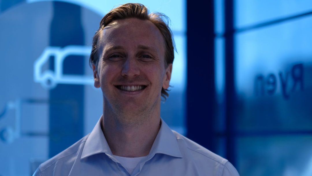 En mann i blå skjorte smiler foran en blå energistasjon.