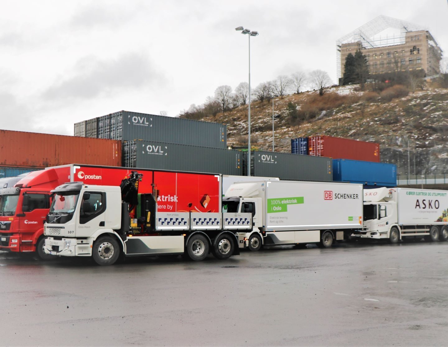 Flere lastebiler parkerte på rekke og rad nær stablede skipscontainere og en høyde med en bygning på toppen. Bakken er våt og overskyet himmel tyder på nylig regn.