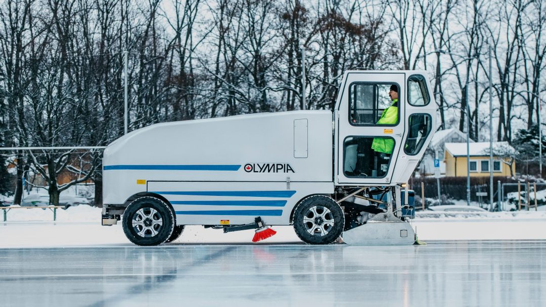En person som kjører en isoppussingsmaskin over en utendørs bane, omgitt av nakne trær og snø, er oppmerksom på kommunale utslipp da de effektivt vedlikeholder den uberørte overflaten.