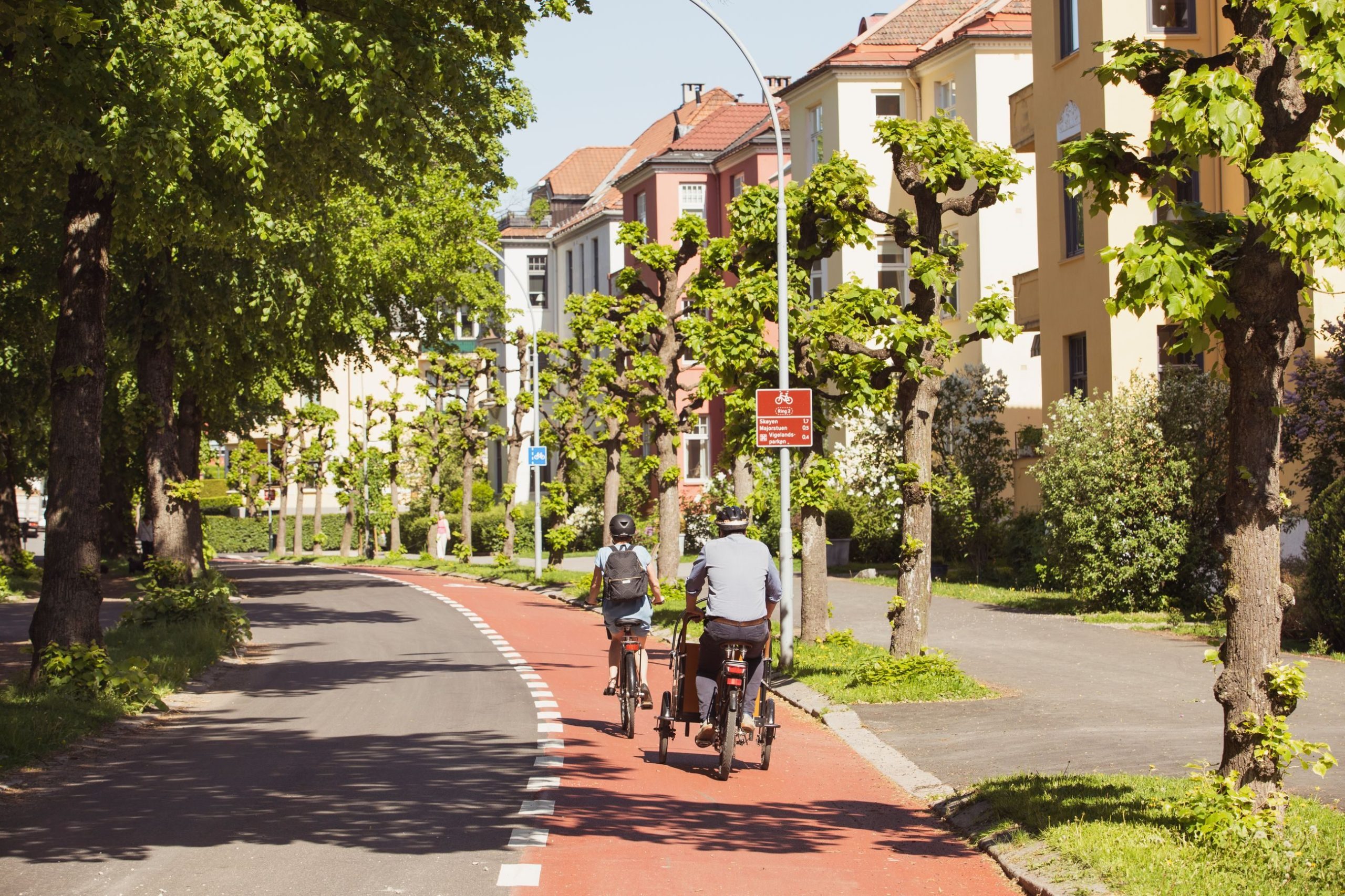 To personer sykler på en utpekt sykkelveier omkranset av trimmede trær, nær bolighus under klar himmel.
