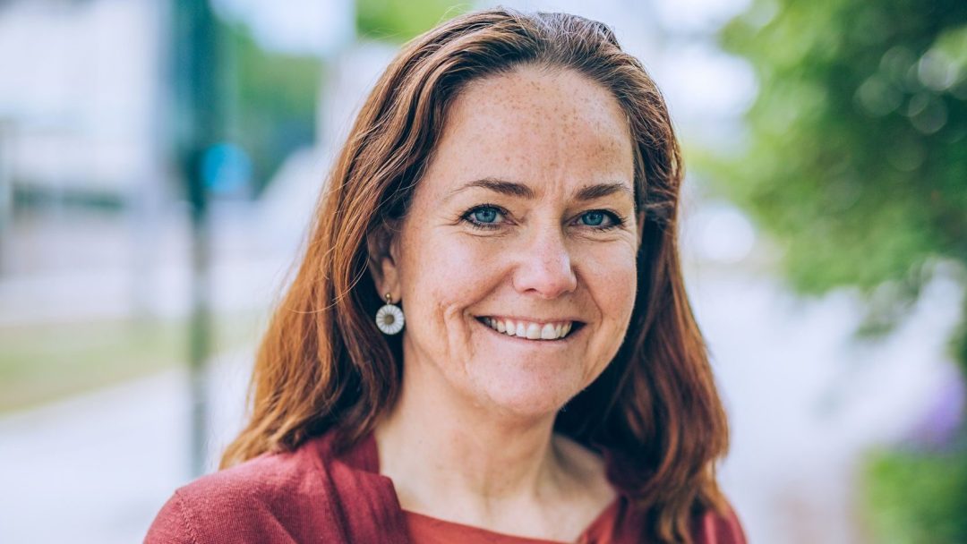 En kvinne med langt rødt hår smiler utendørs og utstråler varme i en rød topp og øredobber. Det uskarpe grøntområdet og en svingete sti bak henne minner en om naturens energiordninger, og fanger harmoni og vitalitet i rolige omgivelser.