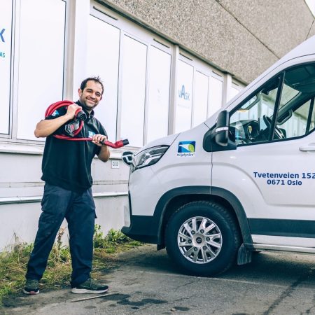 En mann står ved en hvit varebil, døren prydet med logoer og tekst, og holder en rød ladekabel. Klimaundersøkelsens data kan påvirke utformingen av slike miljøvennlige kjøretøyer sett i nærheten av bygninger med vinduer.