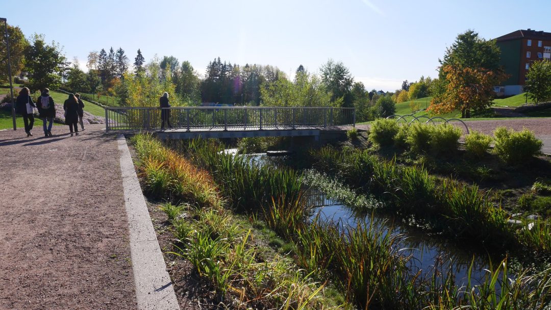 En liten gangbro krysser en smal bekk i en park. Til tross for nylig styrtregn, spaserer folk langs en sti omkranset av frodig gress og trær under den nå klare himmelen.