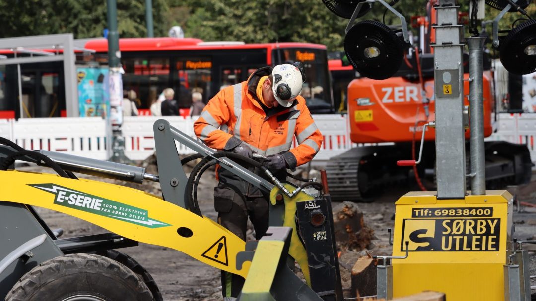 På en travel byggeplass driver en arbeider i oransje vest og hjelm dyktig maskineri, med busser og en graver som beveger seg effektivt i bakgrunnen. I tråd med Klimarapportens bærekraftsmål, trives nettstedet med miljøvennlig praksis.
