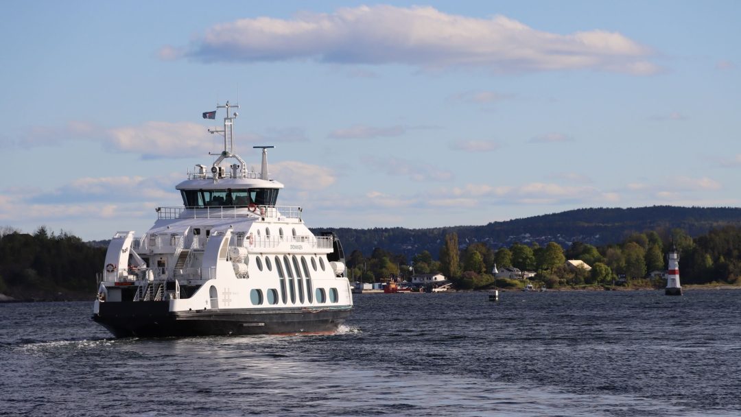 En hvit ferge glir over Oslo havn, satt i bakgrunn av kupert strandlinje og spredte bygninger under en delvis overskyet himmel.