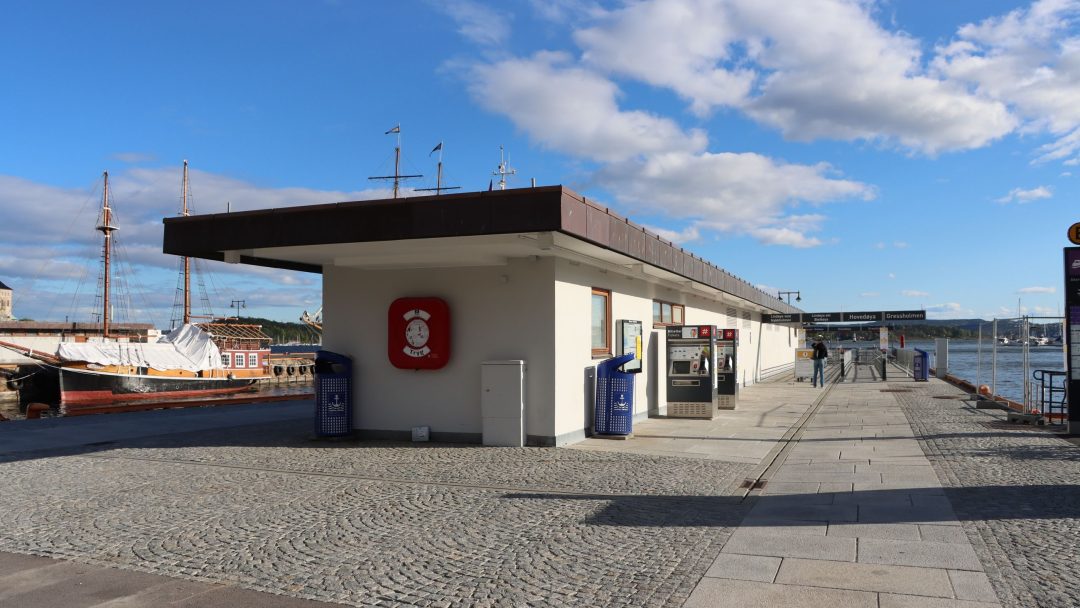 Et billettkontor ved sjøen i Oslo havn har salgsautomater og brannalarm, med et skip til kai i nærheten. Brosteinsganger strekker seg under en delvis overskyet himmel.