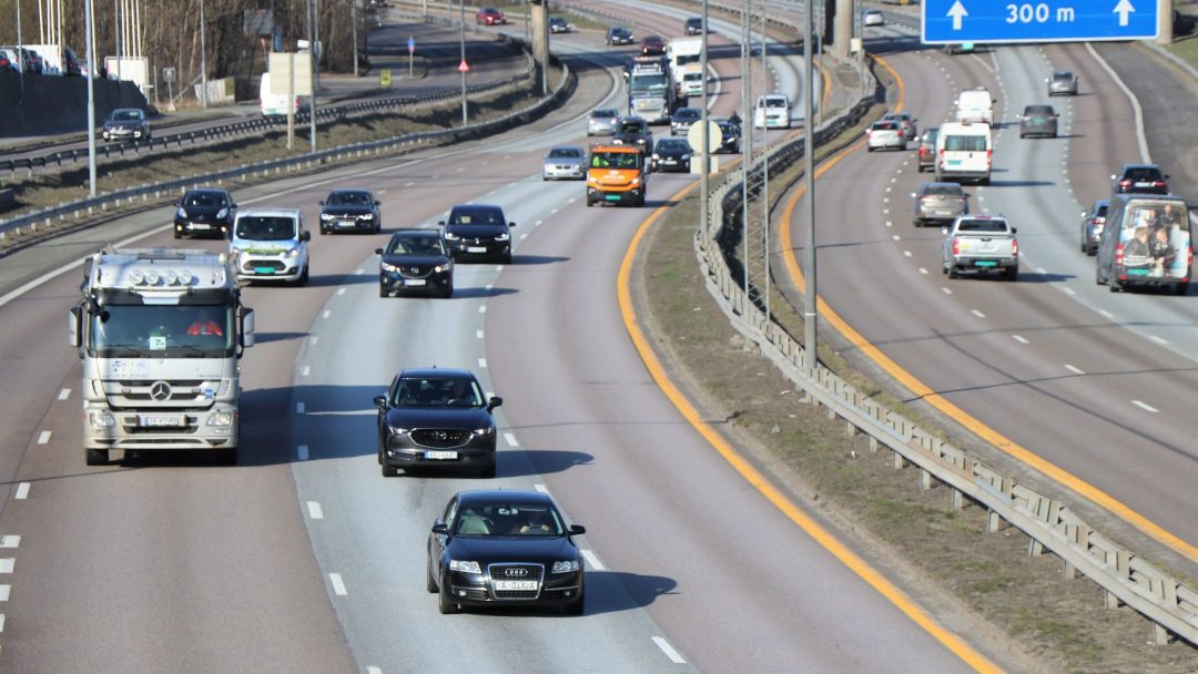 Biler og lastebiler navigerer en flerfelts motorvei under klar himmel, med tanke på deres innvirkning på klimaet. Et blått retningsskilt som indikerer 300 meter er synlig til høyre, og minner sjåførene om avstanden deres når de vurderer drivstofforbruket innenfor klimabudsjettene.