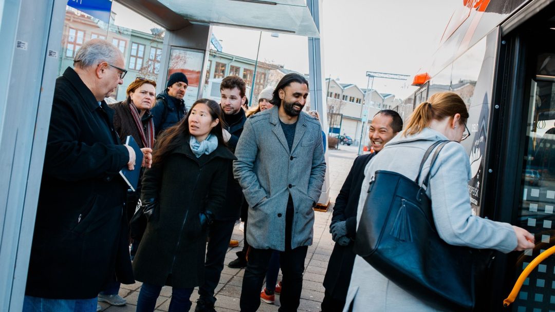 En gruppe mennesker venter på en bussholdeplass, noen iført frakker mens de omfavner kulden fra miljøhovedstadsåret. En kvinne går om bord i bussen mens andre står i nærheten, omgitt av bygninger i bakgrunnen.