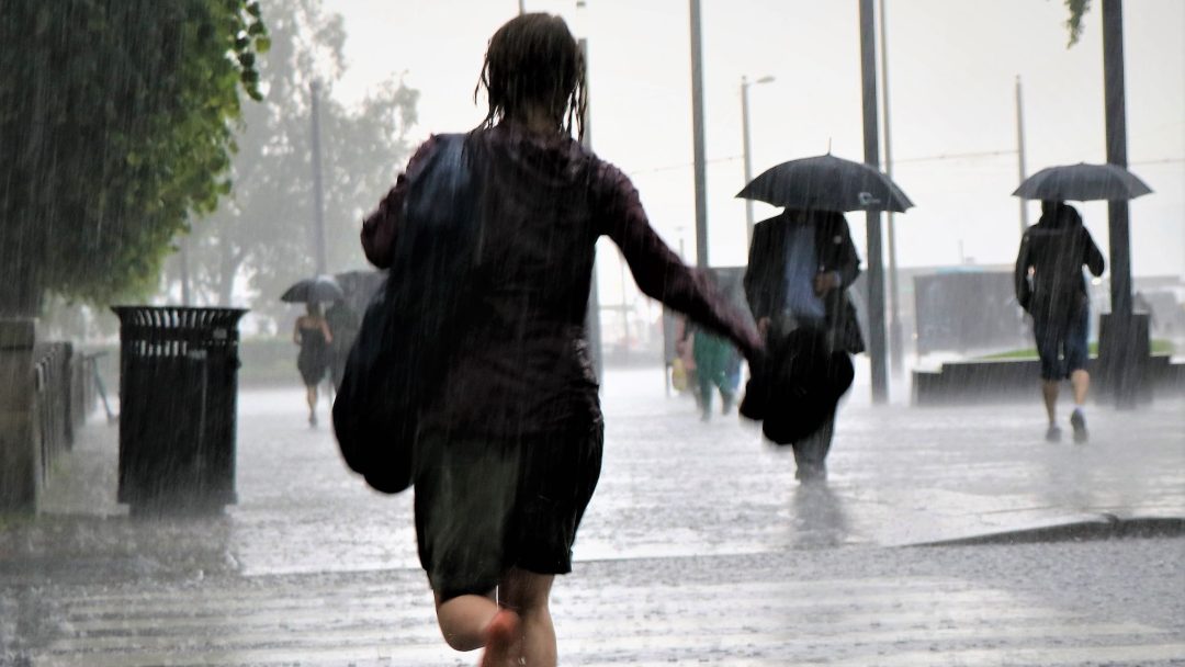 Løper i regnet med vått hår og klær, holder Klimarapporten i den ene hånden mens andre rusler forbi under paraplyene. Scenen er et grått, vått vitnesbyrd om motstandskraft midt i dystert vær.