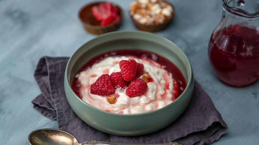 En skål med yoghurt toppet med bringebær står på et mørkt klede, som minner om grøtrester. I nærheten ligger en skje, to boller fylt med nøtter og bær, og en glassmugge med rød væske.