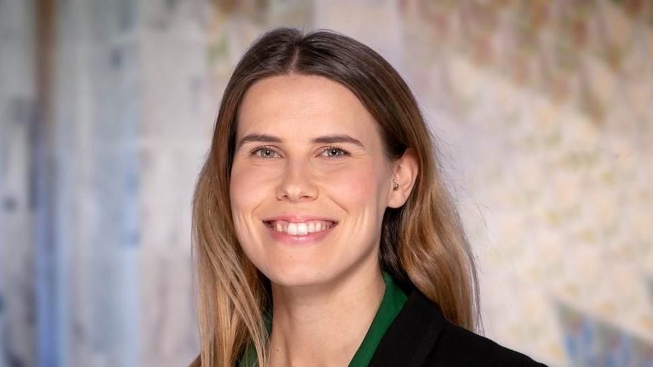 Kvinne med langt hår som smiler, iført svart jakke over en grønn topp, stående foran en mykt uskarp bakgrunn, og utstråler selvtilliten som sømmer seg for et Oslo Handelsstands Forening-medlem.