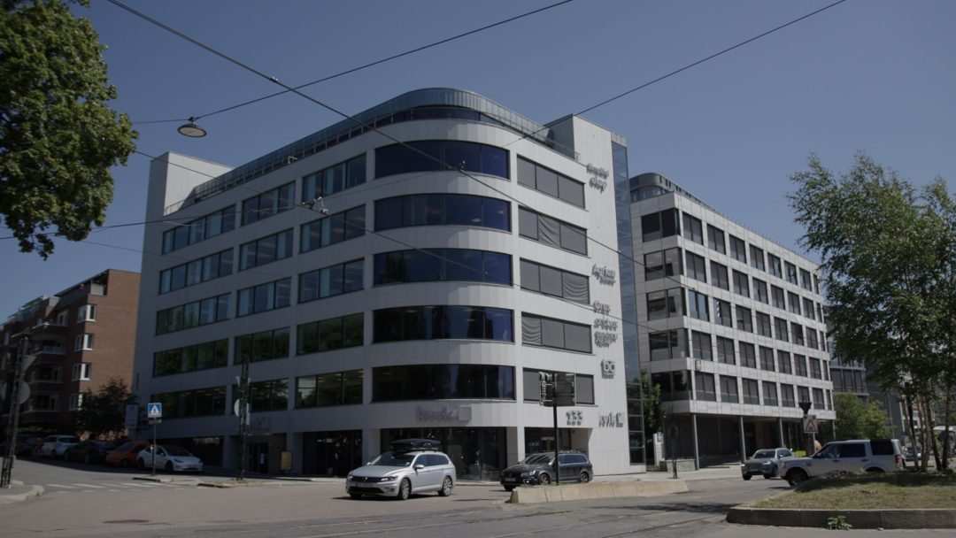 Det store moderne kontorbygget, med et avrundet hjørne og glassvinduer, står under en klarblå himmel. Med biler parkert på gaten og et tre i forgrunnen, utfyller den perfekt det miljøvennlige fokuset inspirert av solcelletilskuddets initiativ.