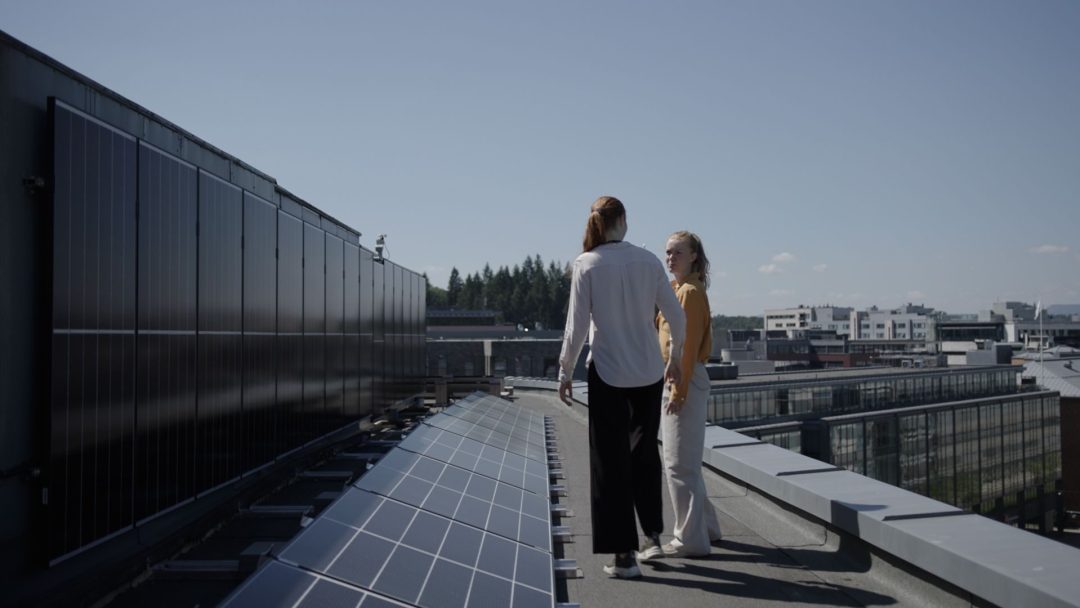 To personer står på et tak i nærheten av solcellepaneler, med utsikt over et bybilde under en klar blå himmel, og betrakter fordelene med solcelletilskuddet.