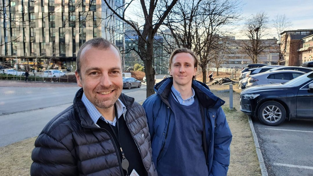 To menn i jakker smilende utendørs nær et tungtransportkjøretøy og en bygning, med nakne trær i bakgrunnen.