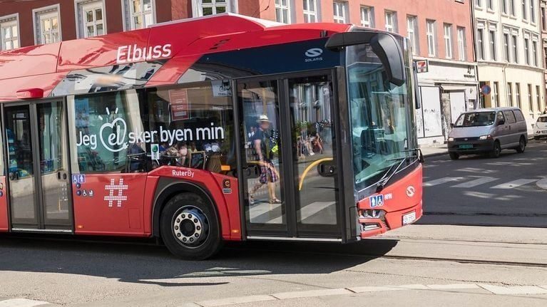 En rød elektrisk buss glir gjennom bygaten, passasjerer synlige gjennom vinduene. Dansk tekst på siden nikker til den siste klimaundersøkelsen, som fremhever byens engasjement for bærekraftig transitt.