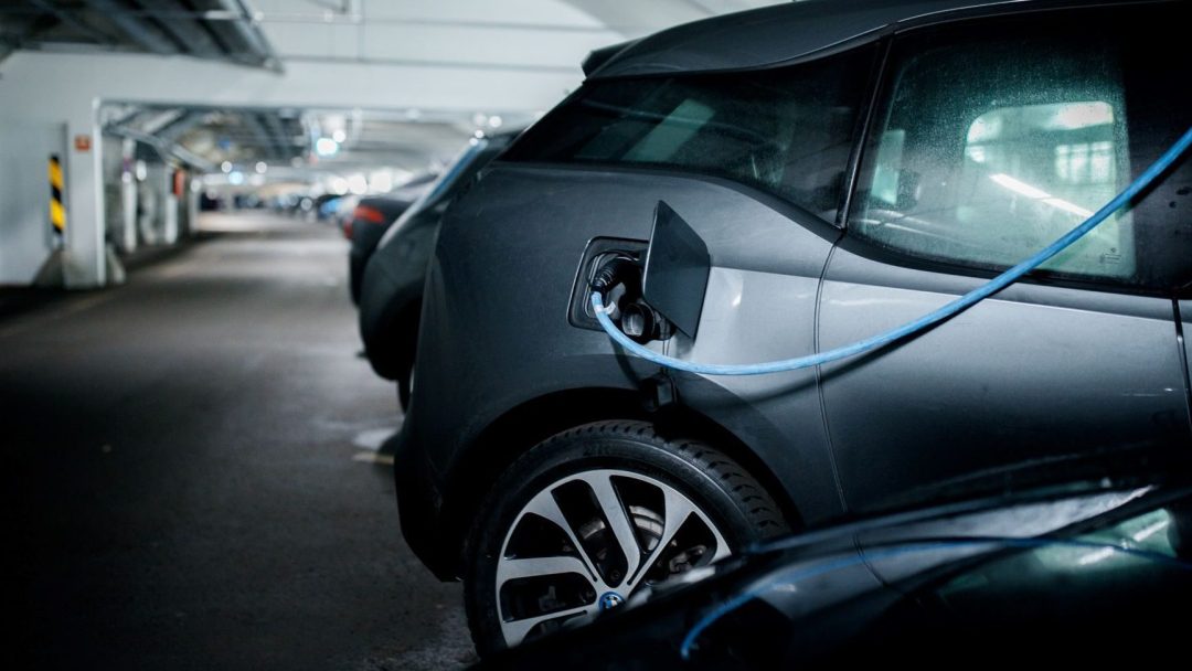 En elbil lader i et parkeringshus, koblet til strømkabel, og bidrar til redusert klimagassregnskap. Andre biler står parkert i bakgrunnen.