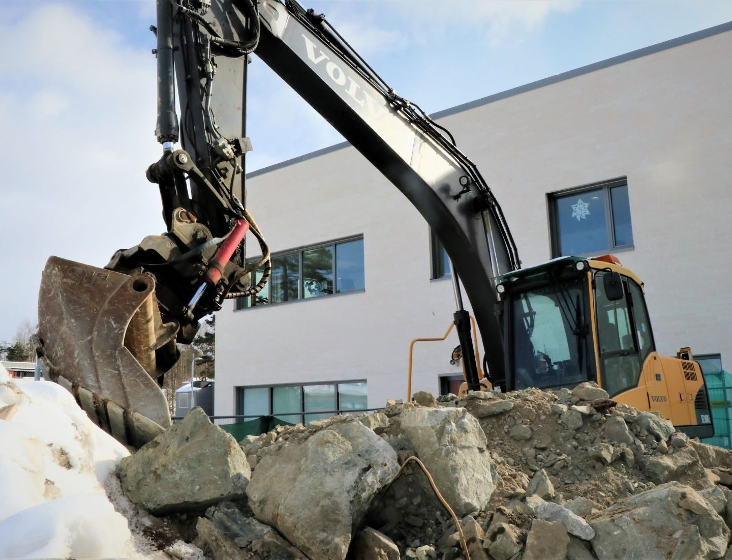 En gravemaskin flytter flittig stein på en byggeplass nær en moderne bygning, og tilpasser driften til prinsippene i Klimabudsjettet for å sikre en miljømessig bærekraftig utvikling.