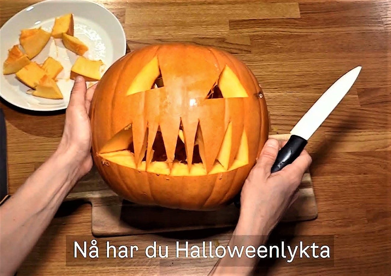 To hender skjærer ut en jack-o'-lanterne fra en grresskar med en kniv på en treflate. Gresskarbiter og en tallerken er i bakgrunnen. Svensk tekst lyder Nå har du Halloweenlykta (Nå har du en Halloween-lykt).