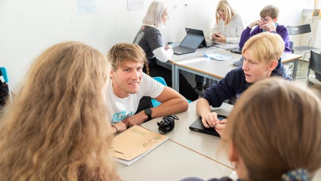 En gruppe mennesker som sitter ved pulter, er engasjert i samtaler og bruker bærbare datamaskiner i klasserom. Hennes booker du en klimapilot, veileder diskusjoner om bærekraft med innovativ innsikt.