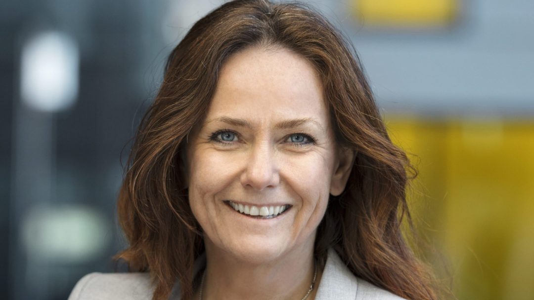 En kvinne med langt brunt hår smiler selvsikkert og tar på seg en lys blazer. Den uskarpe bakgrunnen har subtile hint av blått og gult, som fremkaller essensen av en klimagassregnskap-atmosfære.