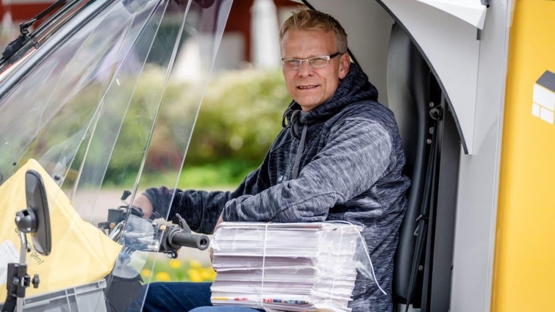 En person med briller og hettegenser sitter på et scooter-lignende kjøretøy og griper en haug med post, og leverer kanskje resultater fra Klimaundersøkelsen.