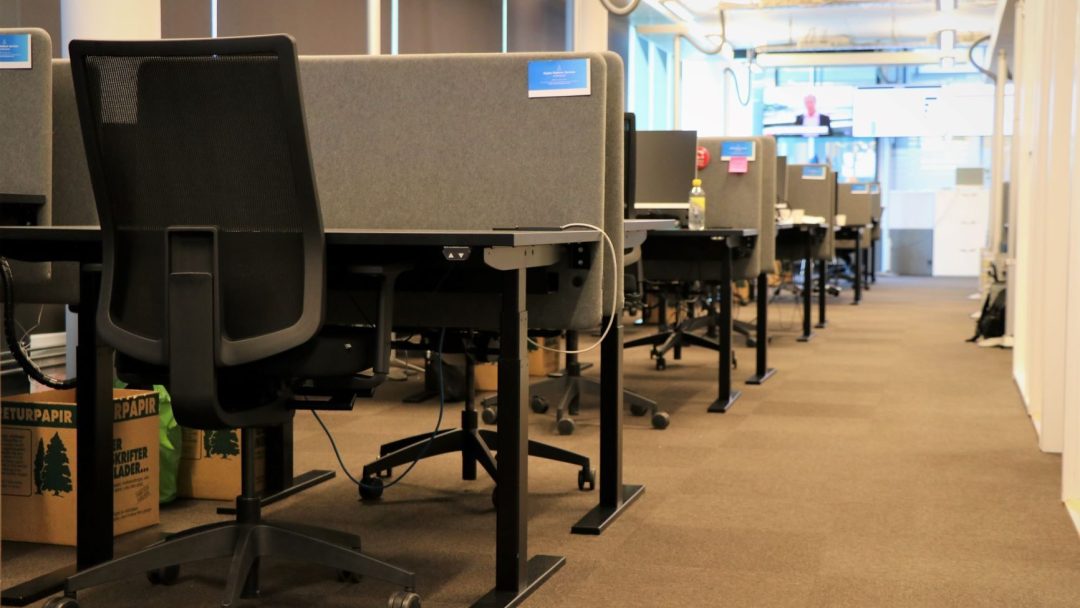 Tom kontorplass med rader med grå avlukker og svarte kontorstoler på et brunt teppebelagt gulv fremkaller en profesjonell, men moderne hjemmekontorstemning. Bokser er synlige under noen pulter, og store vinduer langs veggen, som inviterer til naturlig lys.