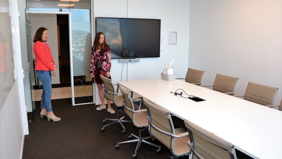 To personer står i et konferanserom med et langbord, svingbare stoler og en stor TV-skjerm på veggen, og blander elementer fra et hjemmekontor for å skape en avslappet, men profesjonell atmosfære.