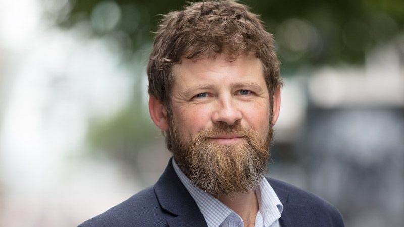 En mann med skjegg og krøllete hår, iført mørk jakke og lys skjorte, står utendørs mot det uskarpe grøntområdet. Hans gjennomtenkte uttrykk tyder på at han kanskje reflekterer over Klimarapportens innsikt i naturen.