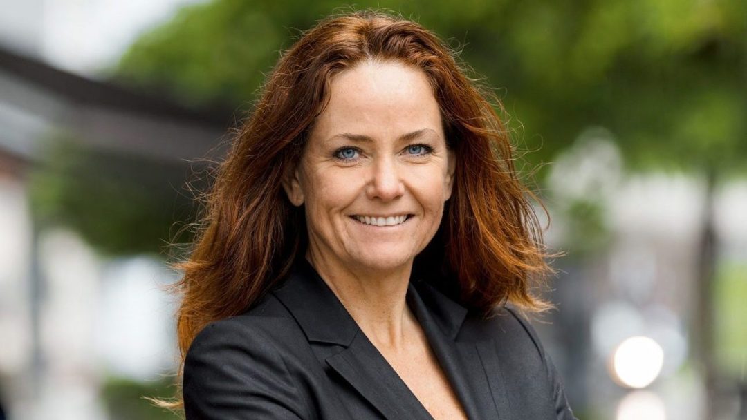 En person med langt rødbrunt hår iført mørk blazer smiler utendørs, det uskarpe grøntområdet bak dem minner subtilt om innsiktene fra klimaundersøkelsen.