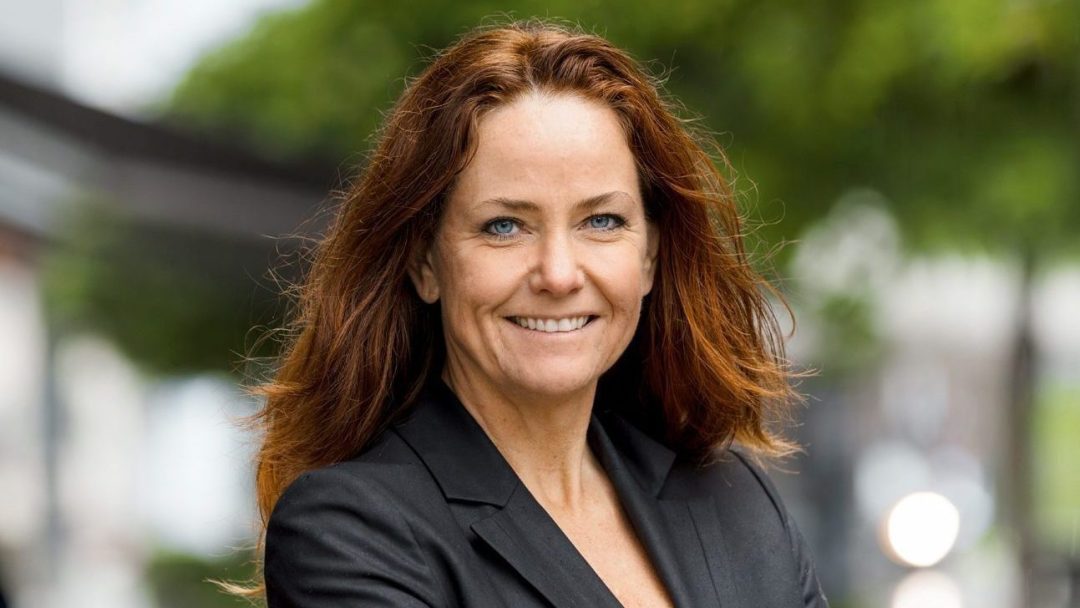 En kvinne med langt rødt hår, iført svart blazer, står utenfor Oslo Handelsstands Forening-bygget og smiler varmt.