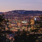 Bybildeutsikt i skumringen, med opplyste bygninger og tresilhuetter i forgrunnen. Bakker og spredt lys er synlige i bakgrunnen under en klar himmel.